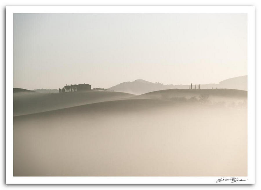 Paesaggio collinare toscano avvolto nella nebbia con una casa e alberi sullo sfondo.