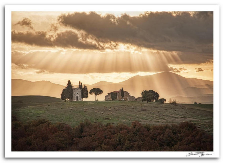 Paesaggio collinare toscano con una chiesetta e un casale illuminati da raggi di luce dorata  tra le nuvole.